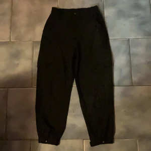 Svarta cargopants med fickor och knappar - Svarta cargopants med flera stora fickor och snygga knappdetaljer vid benslut och bak. Byxorna har elastisk midja och är tillverkade i ett mjukt bomullstyg. Perfekta för en chill och avslappnad stil. Passar även S.