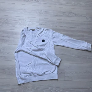 Vit sweatshirt från Moncler med tryck - Vit långärmad sweatshirt från Moncler med svart logomärke på bröstet och stor svart text på ryggen. Tröjan har rund halsringning och är tillverkad i mjuk bomull. Perfekt för dig som gillar stilrena och exklusiva plagg med streetkänsla.