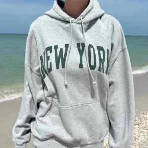 Brandy Melville Hoodie - Säljer en oversized grå hoodie med mörkgrönt New York-tryck på bröstet. Tröjan har huva med snörning och en stor magficka. Perfekt för chill dagar och streetstyle. Mjuk insida och långa ärmar för extra comfort.