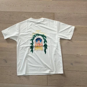 Vit Casablanca t-shirt med tryck - Vit t-shirt från Casablanca med färgglatt tryck på ryggen som föreställer en byggnad och gröna blad. T-shirten har klassisk passform och korta ärmar. Perfekt för dig som gillar unika prints och stilren design.