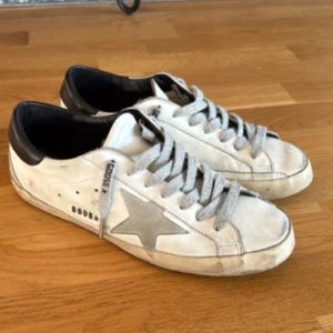 Golden Goose Superstar sneakers vit/grå - Golden Goose sneakers i vitt skinn med grå stjärna på sidan. Köpta i USA, hälpartiet är svart. Superbra skick har bara använts några gånger. Skorna levereras med originalpåse och tillbehör. 