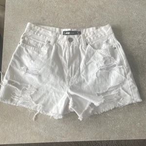 Vita slitna jeansshorts från FB Sister - Vita jeansshorts från FB Sister med hög midja, råa kanter och slitna detaljer framtill och baktill. Klassisk femficksmodell i denim med knapp och dragkedja. Perfekta för sommaren och ger en cool, avslappnad vibe.