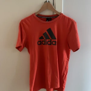 Orangeröd Adidas t-shirt med logga - Röd t-shirt från Adidas med klassisk svart logga på bröstet. T-shirten har rund halsringning och korta ärmar. Perfekt för dig som gillar sportig stil och vill ha något bekvämt och snyggt till vardagen.