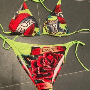  bikini med tatueringsmönster - Färgstark bikini  med ikoniskt tatueringsinspirerat mönster, röda rosor och text. Triangeltopp och trosa med knyt i sidorna, detaljer i limegrönt. Perfekt för dig som vill sticka ut på stranden.
