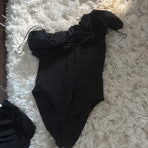 Body - Säljer en svart oneshoulder body från Anna Field med volangdetalj längs axeln. Perfekt för dig som vill sticka ut med en cool och trendig look.