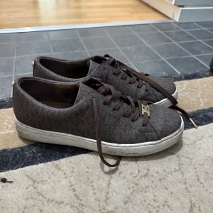 Bruna sneakers från Michael Kors - Snygga bruna sneakers från Michael Kors med monogrammönster och vita sulor. Skorna har bruna snören och guldfärgade MK-detaljer framtill. Tillverkade i syntetmaterial med rund tå och klassisk sneakerstil. Perfekta för dig som gillar stilrena och trendiga skor.