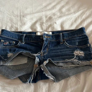 Mörkblå low waisted jeansshorts från Hollister - Säljer ett par mörkblå jeansshorts från Hollister med slitna detaljer och fransig kant. Klassisk femficksmodell med snygga sömmar på bakfickorna. Perfekta för sommaren och har låg midja.