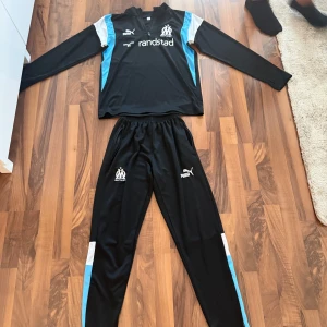 Marseille dress - Svart dress från Puma med Olympique de Marseille-logga på ena benet och Puma-logga på andra. Byxorna har elastisk midja, dragkedjefickor och detaljer i vitt och ljusblått längs benen. Perfekta för fotboll eller träning.