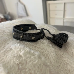 Christian dior liknande armband  - Snyggt grått armband i tyg med små silvriga stjärnor längs bandet. Armbandet har knytning med två tofsar i ändarna. Perfekt accessoar för att lyfta din outfit med lite extra edge.