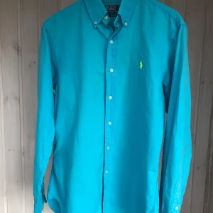 Turkos skjorta från Polo Ralph Lauren - Snygg turkos skjorta från Polo Ralph Lauren i slim fit-modell. Skjortan har klassisk krage med knappar, lång ärm och ikonisk broderad logga i neongrönt på bröstet. Tillverkad i bomull och perfekt för dig som gillar stilrena och färgstarka plagg. Slitage finns vid kragen, därav priset, inget som ska synas när man har på sig skjortan.