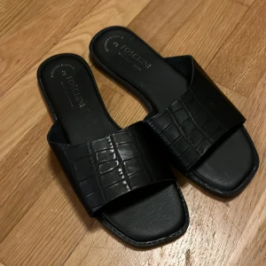 Svarta sandaler från Foschini - Stilrena svarta sandaler från Toscanini med bred rem i croco-mönstrat skinn. Snygg fyrkantig tå och platt sula gör dem perfekta för en chill look. Passar till allt och är superenkla att glida i.
