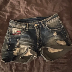  Low waist Jeansshorts med stjärnor och slitningar - Snygga blå jeansshorts med råa kanter och slitna detaljer. Bakfickan har broderade stjärnor och röda ränder som ger en cool, amerikansk vibe. Klassisk femficksmodell med knapp och dragkedja. Perfekta för sommaren och festivaler.
