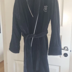 Svart badrock från Gant Home L - Säljer en svart badrock från Gant Home i storlek L. Badrocken är i mjuk frotté, har vit piping längs kanterna, broderad logga på bröstet och knytskärp i midjan. Perfekt för chill efter duschen eller morgonmyset.