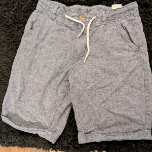 Grå blå linne-shorts med snörning stl 146 - Snygga grå shorts i linne med avslappnad passform och vita snören i midjan. De har fickor på sidorna, knapp och uppvikta benslut. Perfekta för varma dagar och ger en chill vibe till din outfit.