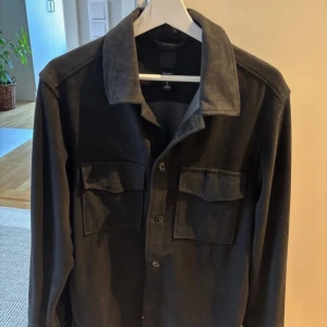 Svart overshirt från H&M, storlek M - Svart overshirt från H&M i regular fit med klassisk krage och två stora bröstfickor med lock. Skjortan har knäppning framtill och långa ärmar. Perfekt att slänga över en t-shirt för en chill look.