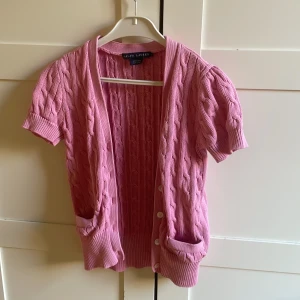 Rosa kabelstickad kofta Ralph Lauren - Supersöt rosa kabelstickad kofta från Ralph Lauren i 100% bomull. Koftan har korta ärmar, djupa fickor framtill och knappar hela vägen. Perfekt för lager-på-lager och ger en preppy vibe till din outfit.