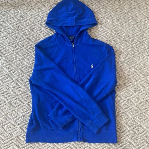 Blå hoodie från Polo Ralph Lauren - Storlek L i barn så S. Snygg blå hoodie från Polo Ralph Lauren med dragkedja och huva. Klassisk design med liten gul broderad logga på bröstet. Mjuk bomullskänsla och långa ärmar, perfekt för chill dagar eller när du vill ha en sportig look.