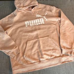 Rosa/aprikos hoodie från Puma med logga - Säljer en snygg rosa/aprikos melerad hoodie från Puma med stor vit logga framtill. Tröjan har huva med dragsko, magficka och ribbade muddar vid ärmslut och nederkant. Aldrig använd men provad o lappar borta.