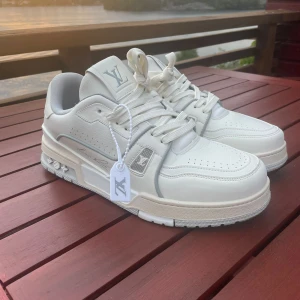 Louis Vuitton Trainers MED KVITTO - Exklusiva Louis Vuitton sneakers i vitt skinn med klassisk LV-logga på plös och sidor. Snygga detaljer på sulan och perforeringar på tån. Skorna har rund tå, snörning och en chunky sula för en modern look. Perfekta för dig som vill sticka ut med lyxig streetstyle.