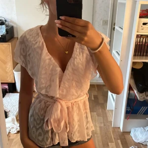 Ljusrosa blus med knyt och volang - Supersöt ljusrosa blus med leopardmönster och omlottkänsla. Den har korta ärmar, volangdetaljer och ett knytband i midjan som ger en snygg siluett. Perfekt för dig som gillar romantiska och trendiga plagg.