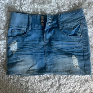 Blå jeanskjol från Dolce Rosa - Snygg blå jeanskjol från Dolce Rosa med slitna detaljer och tre diamant liknande knappar framtill. Klassisk femficksmodell och kort passform som sitter tajt. Perfekt för dig som gillar en vintage och trendig look.