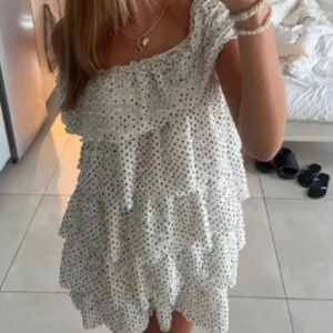 Vit volangklänning med prickar - Supersöt kort volangklänning i vitt med svarta prickar. Offshoulder-modell med flera lager volanger som ger en fluffig och lekfull look. Perfekt för varma dagar och sommarhäng. Lätt och luftig känsla, passar dig som gillar en trendig och söt stil.