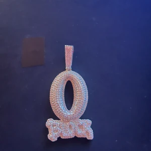 Iced out silverhalsband med O-block - Säljer ett halsband i silverfärg med en stor, glittrig O-formad berlock. Berlocken är täckt av små stenar som ger en riktigt blingad känsla och har texten 'BLOCK' i botten. Kedjan är tvinnad och matchar berlocken perfekt för en cool streetstyle.