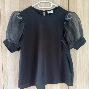 Svart blus med puffärmar från Aware - Supersnygg svart blus från Aware by Vero Moda med transparenta puffärmar i organza och rund halsringning. Korta ärmar med bred mudd ger en cool siluett. Perfekt för dig som vill sticka ut lite extra med en trendig och stilren topp.