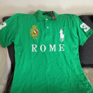 Grön Polo Ralph Lauren t-shirt Rome - Snygg grön t-shirt från Polo Ralph Lauren med vit broderad logga och texten ROME på bröstet. Klassisk pikékrage, korta ärmar och broderad detalj med Italiens emblem. Perfekt för dig som gillar sportig och preppy stil.