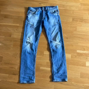 Blå slitna jeans med hål - Säljer ett par blå jeans med slitningar och hål på båda benen. De har klassisk femficksdesign och raka ben. Perfekta för en avslappnad och trendig look.