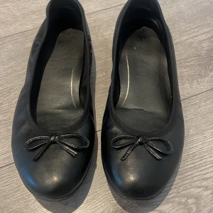 Svarta ballerinaskor med rosett - Klassiska svarta ballerinaskor i skinn med rund tå och söt rosettdetalj framtill. Perfekta för en stilren och enkel look, passar till både jeans och klänning. 