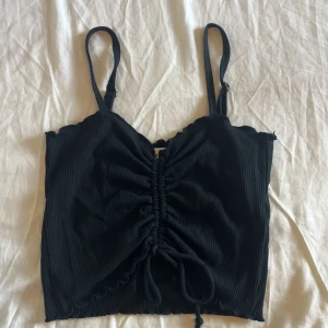 Svart ribbad linnetopp med snörning - Svart ribbad linnetopp med justerbara smala axelband och snygg snörning framtill som ger en rynkad effekt. Toppen är croppad och har en enkel, stilren look som passar perfekt till jeans eller kjol. Materialet är mjukt och stretchigt för en bekväm passform.