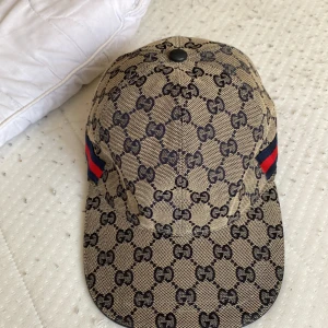 Gucci keps med GG-mönster och röd-blå detalj - Snygg keps från Gucci i beige med klassiskt GG-monogram över hela ytan. Kepsen har en böjd skärm och ikoniska röd-blå ränder på sidan. Tillverkad i bomullsmix med mörkblå insida. Perfekt för dig som vill sticka ut med lyxig streetstyle. Kommer med box