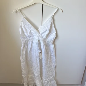 Vit somrig bomullsklänning med brodyr - Superfin vit långklänning i 100% bomull med broderade detaljer och volangkant nertill. Klänningen har smala axelband, omlott-v-ringning och knytband med tofsar i midjan. Perfekt för varma dagar och har en lätt och luftig känsla. Den passar  s xs men kan även passa fler storlekar för den är stretchig och axelbanden går att spänna åt🩷