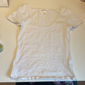 Vit urringad t-shirt från H&M - Enkel och fräsch vit t-shirt från H&M i mjuk bomull. Modellen har korta ärmar och en djup rundringning. 