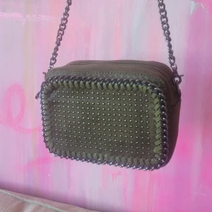 Militärgrön väska med kedja - Snygg militärgrön väska i mocka med coola silverfärgade nitar på framsidan och grov kedjerem. Väskan har rektangulär form, dragkedja upptill och dekorativ kant. Perfekt för dig som gillar edgy accessoarer. Så himla snygg men kommer inte till användning! Aldrig använd!!💚