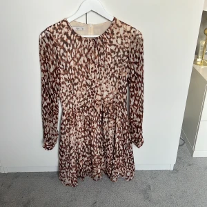 Brun leopardmönstrad klänning MNG - Kort klänning från MNG med brunt och beige leopardmönster. Långärmad modell med rund halsringning och dragkedja bak. Tunt, lätt material som ger ett flowy intryck. Perfekt för dig som gillar djurmönster och vill sticka ut.