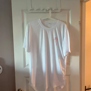 Vit t-shirt med ros-tryck - Vit t-shirt i bomull med korta ärmar och rund hals. På bröstet finns ett litet tryck av en rosa ros med droppar. Enkel och clean design som passar till det mesta. Skön passform och lite längre modell.