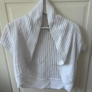 Vit stickad kofta från H&M - Säljer en vit stickad kofta från H&M med korta ärmar och öppen front. Koftan har ett snyggt hålmönster och bred ribbad krage som ger en chill och trendig vibe. Perfekt att slänga över en topp under vår eller sommarkvällar.