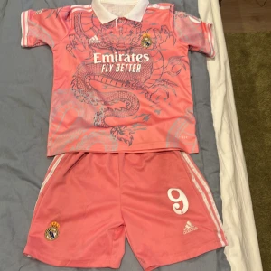 Real Madrid Mbappé rosa fotbollströja set - Säljer ett fett Real Madrid fotbollskit i rosa från Adidas, med Mbappé och nummer 9 på ryggen. Tröjan har korta ärmar, vit krage och drakmotiv i grått. Broderade märken, Champions League-patch och klassiska Adidas-ränder på shortsen. Materialet är lätt och andas.