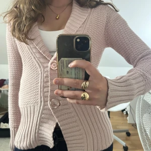 Ljusrosa stickad kofta med stora knappar - Superfin ljusrosa stickad kofta med två stora knappar framtill och bred ribbad kant. Koftan har lång ärm och en mysig, lite oversized krage. Perfekt att slänga på sig över ett linne eller t-shirt för en chill look.