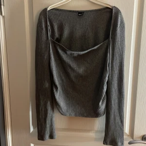 Grå ribbad topp från Gina Tricot - Säljer en grå ribbad långärmad topp från Gina Tricot i storlek XL. Toppen har en snygg draperad urringning och är tillverkad i ett mjukt, stretchigt material. Perfekt för dig som gillar stilrena och bekväma plagg med lite edge.
