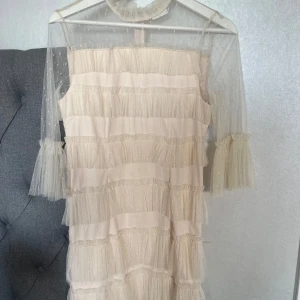 Ljusbeige volangklänning med spets/tyll - Superfin ljusbeige kortklänning med volanger och transparenta ärmar i prickig tyll. Klänningen har hög krage med volang och trekvartsärm med extra volangdetalj. Perfekt för dig som gillar romantisk och söt stil.