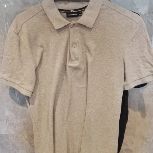 Grå pikétröja från J.Lindeberg - Snygg ljusgrå pikétröja från J.Lindeberg med klassisk krage och två knappar framtill. Tröjan är i mjuk bomull och har korta ärmar. Perfekt för en clean och stilren look, med diskret logga på bröstet.