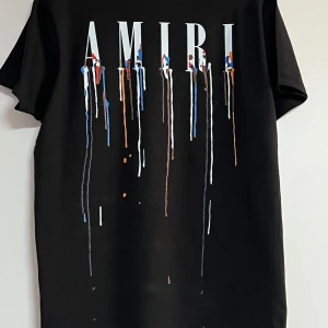 Svart AMIRI t-shirt med färgstänk - Svart t-shirt från AMIRI med stort tryck framtill. Trycket består av AMIRI-loggan och färgstänk i blått, vitt, rött och orange som rinner ner över bröstet. Klassisk passform och korta ärmar. Perfekt statement-plagg för dig som gillar streetstyle.