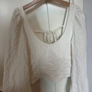 Vit  blus med knyt i ryggen - Superfin vit croppad blus med puffiga långärmar och struktur i tyget. Toppen har en djup urringning och snygg knytning i ryggen.  