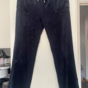 Mörkblå velourbyxor Juicy Couture - Mörkblå mjukisbyxor från Juicy Couture i mjuk velour. Klassisk logga broderad på ena fickan och elastisk midja med snörning. Perfekta för en chill och trendig look. Har en liten fläck som jag visa på bilden, man ser inte den så mycket, skriv vid intresse eller mer info