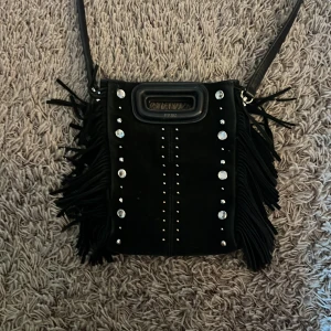 Svart mocka axelväska med fransar från Maje - Cool svart axelväska i mocka från Maje med långa fransar på sidorna och silverfärgade nitar och stenar framtill. Väskan har ett robust handtag upptill och justerbar axelrem. Perfekt för dig som vill sticka ut med detaljer och edgy stil.