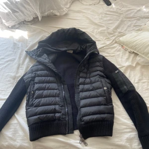 Svart Moncler cardigan - Säljer en svart cardigan från Moncler med stickade ärmar och huva. Jackan har quiltad front och rygg i glansig polyester, medan ärmarna är grovstickade. Dragkedja framtill och fickor med tryckknapp. Klassisk Moncler-logga på ena ärmen.