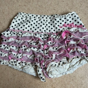 Prickiga volangshorts Angel Blue - Supergulliga vita shorts med svarta prickar från Angel Blue. Shortsen har flera lager volanger med rosa glitterdetaljer och elastisk, de är mini shorts. 